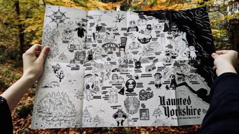 A person holds a map titled Haunted Yorkshire in a forest. The map features playful illustrations of aliens, giants, fairies and ghosts attached to location names. 