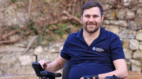 A man sitting in a wheelchair wearing a blue polo top with an RAF logo on it. He is smiling and there is a bush and a stone wall behind him.