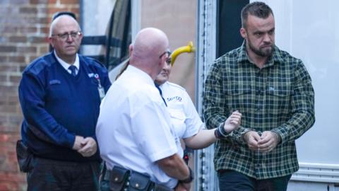 Robbie Cook, wearing a green check shirt and blue jeans, in handcuffs, being lead out of a prison van and into Lewes Crown Court by three uniformed security officers.