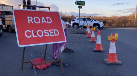 A red 'road closed' sign by a roundabout. Four orange cones are in front of the sign and cars are queuing at the roundabout on the opposite carriageway