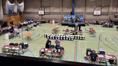 Image of an election count in Huntingdonshire. It is in a sports hall with ballot boxes lying around and tables where people are set to council and adjudicate.
