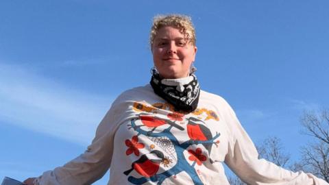 Paradox Mercer looking at camera. She has blonde curly hair. She is wearing a white long-sleeve top with an image of a blue branch with red and dark blue birds on it. There is blue sky in the background with trees. She has a bandanna round her neck. 