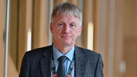 Ivan McKee, a grey-haired man wearing a dark grey suit, blue shirt and dark tie, walking towards the camera in the Scottish Parliament. He is visible from the chest up and has a neutral expression on his face. 