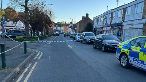 A police cordon in a street with a car park and shops