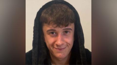 Shay Bladen smiling into the camera. He has brown, short hair and has a navy blue dressing gown hood over his head.