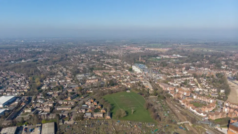 A bird's eye view over Wokingham, including a football pitch, a train line, homes, a factory to the left of the picture, and a church with a steeple in the middle of it