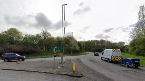 A view of a roundabout showing a white van pulling a trailer and two other vehicles.