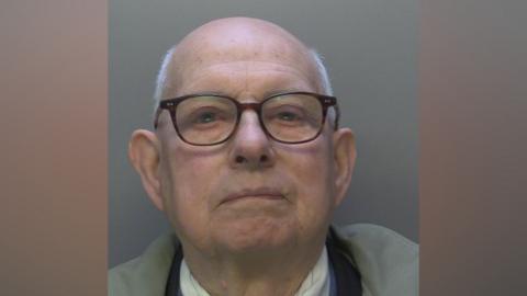 A bald man with short grey hair around the side of his head. He has black glasses.