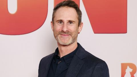 A grey-haired man wearing a dark blue suit smiles at the camera.