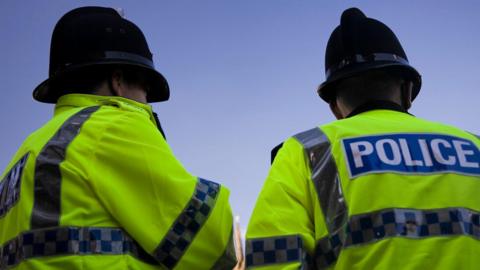 Stock photo of two police officers with their backs to the camera. They are wearing high visibility yellow police jackets and police hats. 