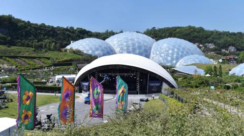 Several bubble-like buildings - known as biomes - at the Eden Project on a sunny day, semi-circular with a stage in front of them. The buildings are set in a now-verdant former quarry  with brightly-coloured flags in front of the stage, with pictures of flowers on them.