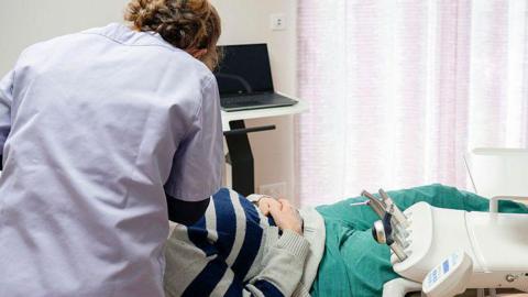 Shows a man in a dentist's chair, wearing a blue and grey striped jumper with green trousers. The female dentist has her back to the camera and is wearing a white dentist's gown.