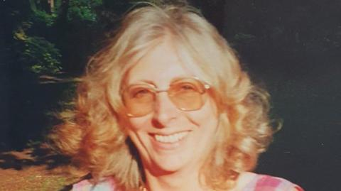 A faded image of a woman with curly blonde shoulder-length hair and tinted sunglasses smiling. Bright sun is shining in her face, and the woodland behind is partially in shadow. She is wearing a checked red and white dress.