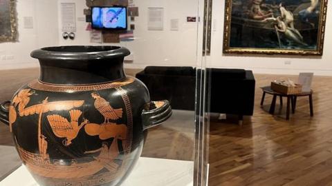 A large black vase featuring an orange-coloured illustration from Greek mythology. It is on display in a large glass case in an art gallery, which paintings in the background.