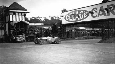A black and white image of a car racing along a concrete track, with people lining the side of it watching.