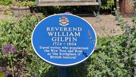 A blue plaque in a garden area with plants and a bench behind it.
