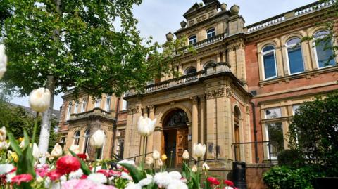 The exterior of Penrith Town Hall. It has an ornate stone, sand-coloured entrance porch which creates a balcony at the first floor. There are colourful flowers, trees and bushes in front of the red stone building.