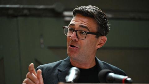 Andy Burnham making a speech at the People's History Museum in Manchester on Monday. He is gesturing with his right hand and is mid-speech. He is wearing glasses, a grey blazer and black shirt.
