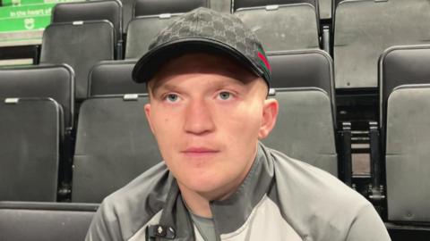A young man wearing a grey tracksuit top and a black cap sits on a fold down black seat in an arena. 