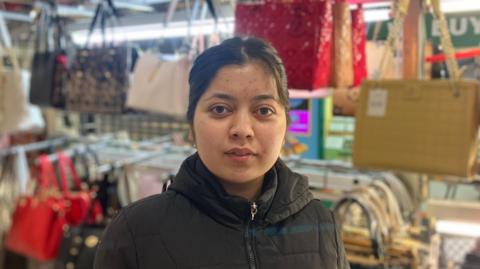 Preet, an Asian woman with dark hair and brown eyes, standing in front of handbags on display on her stall.