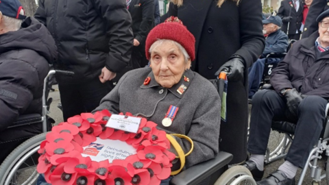 Mary Cliff, 98, wearing red hat with her medal on a grey coat holding a wreath