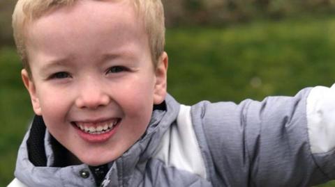 A boy with short blonde hair and a grey coat smiles as he holds out his left arm.