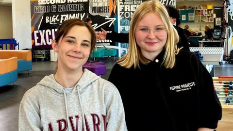 Two girls smiling at the camera as they pose for a picture in a youth club. Maisie on the left has brown hair pulled back in a pony tail and is wearing a grey hoodie with Harvard University branding in large red type. Iga on the right wears a black hoodie bunched up on her arms. She has shoulder-length blond hair and has rimless glasses.