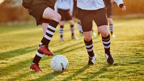 Two young footballers wearing black shorts and black and white striped socks are in the foreground on a pitch - one performs footwork with the ball to avoid being tackled. A number of other players are out of focus in the background.