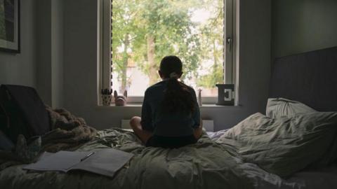 a young girl sitting on a bed looking out of the window