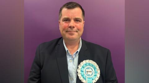 James smiles at the camera standing against a purple backdrop. He wears a black suit jacket and the Reform rosette on his lapel, which is white and pale turquoise.