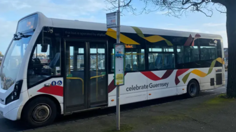A bus parked by a bus stop sign. It is multi-coloured and has the words 'celebrate Guernsey' written on the side.