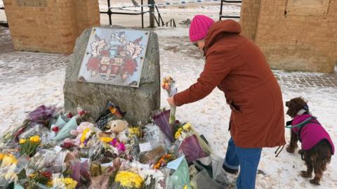 A mount of flowers of various colours lay in the snow in front of two large castellated towers on a seafront. In the background is the sea. A woman wearing a red coat is placing flowers. She has a brown springer spaniel dog on a lead next to her.