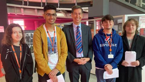 Four students hold their results envelopes and smile along side the principal in the college foyer