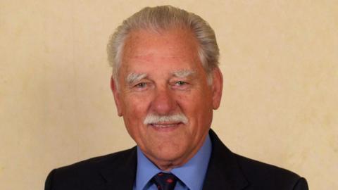A man with grey hair and a moustache is wearing a black suit with a blue shirt and a dark blue tie. He is looking at the camera and is smiling for the photograph. He is standing against a light-coloured backdrop.
