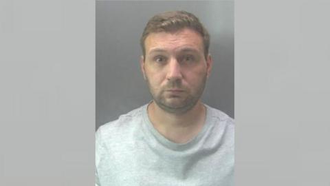 Shaun Fox's mugshot image. He is staring, expressionless at the camera, wearing a grey sweatshirt. He has a stubble beard and brown hair. 