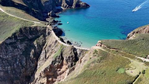 Aerial shot of La Coupée, a narrow road in Sark which connects two parts of the island. The road has a rockface below it. A boat can be seen heading towards the island in the top right-hand corner of the image.