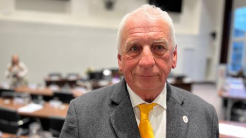 Barry Aspinell wears a grey suit, yellow tie and white shirt. He has white hair. He looks just off camera, with a council chamber out of focus behind him.