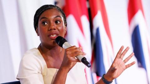 Kemi Badenoch holds a microphone and gestures with her hand. She is sitting in front of a UK flag.