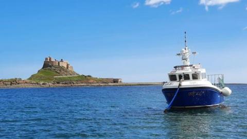 A small fishing boat with a blue hull and white cab and deck is moored up off Holy Island, with a castle on a hill in the background