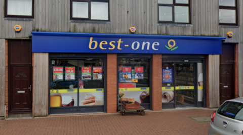A shop with the words Best One on a blue background at the top of the ground floor outside. Part of a pavement and a silver car are visible in front of the store.