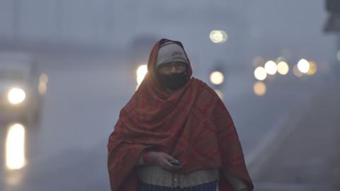 A woman wearing a shawl walks on a road in Uttar Pradesh state, with vehicle lights barely visible in the background because of the dense fog. 