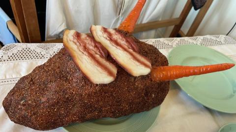 An object made to look like the shape of a turkey with two carrots pointing out of it. It is on a green plate on a white tablecloth.