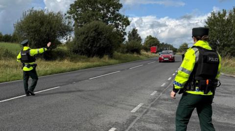 Two police officers are standing at opposite sides of a main road. They are wearing full uniform including hats, high visibility jackets and padded vests.