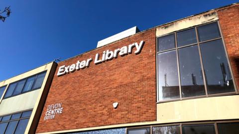 The front of Exeter Library with a large white sign on a brown brick wall and windows to either side.
