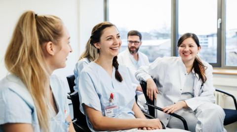 Three women doctors sit on chairs smiling and chatting. 