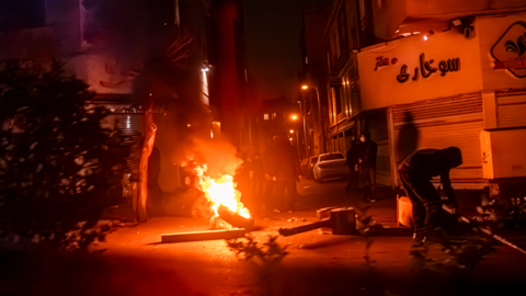 A scene of rioting in Iran - it is a wide shot of an urban street and a fire burns at a small barricade in the middle of the image, making the whole photo an orange-red colour. To the left of the camera is a bush, to the right a figure in black is hunched over a metal object. In the middle distance more black hooded figures can be seen.