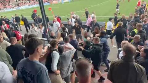 A large group of people in a crowd at Kingsholm rugby stadium start fighting near the pitch, behind the barriers for the stand. The pitch is behind them. Other members of the crowd can be seen watching on.