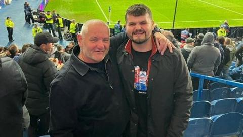 Two men posing for the camera while standing in a seated section of a stadium. The man on the right is bald, wearing a black jacket and has his left arm around the other man's shoulders. The other man has short brown hair, a beard, a brown jacket and black T-shirt with pictures of cans on it.