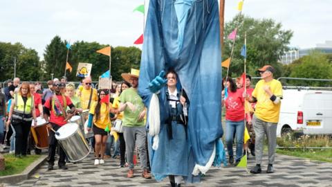 A woman wears a giant blue costume, surrounded by a parade of people in yellow and red tops carrying drums.