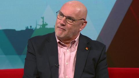 Nick Adams-King is bald and wearing thin circular glasses. He has a dark blazer on over a pink shirt. He is sitting on a red sofa with a red and blue wall of graphics behind him.